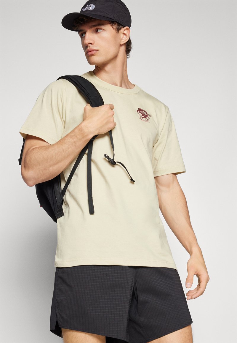 T-shirt en coton beige clair avec un petit graphique bordeaux, associé à un short texturé noir et un sac à dos noir. Design et coupe clairs.