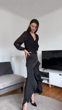 Kvinna poserar inomhus iförd en svart blus med genomskinliga ärmar, en svart asymmetrisk kjol med volanger och svarta högklackade skor.