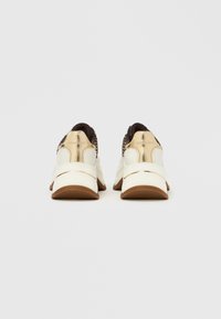 Λευκά και καφέ αθλητικά παπούτσια με κομψό σχεδιασμό, διαθέτοντας υφασμένο άνω μέρος, μεταλλικές λεπτομέρειες και λαστιχένια σόλα για καλύτερη πρόσφυση.