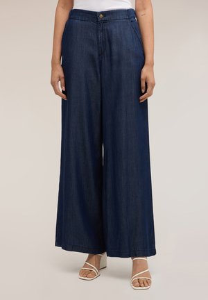 Mujer que lleva pantalones vaqueros azul oscuro de tiro alto y pierna ancha con botón delantero y sandalias blancas de tacón con tiras.