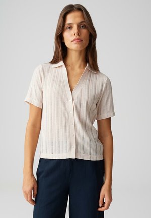 Donna che indossa una camicetta rosa pallido a maniche corte con bottoni e righe verticali, e pantaloni blu scuro, in piedi davanti a uno sfondo uniforme.