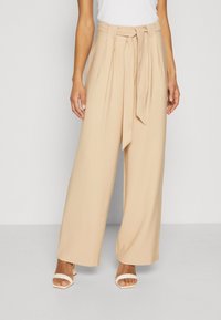 Beige broek met wijde pijpen en een tailleband met strik, gemaakt van gladde stof. Voorzien van plooien aan de voorkant en een ontspannen pasvorm, gecombineerd met witte sandalen met hak.