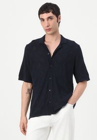Cămașă scurtă cu mânecă, de culoare bleumarin, cu un model texturat în diamante, guler ribbit și nasturi negri, purtată cu pantaloni albi.