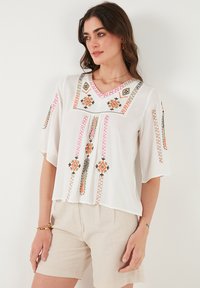 Blusa blanca con mangas cortas, escote en V, con bordados detallados en rosa, naranja y verde. Tejido ligero con un corte holgado.