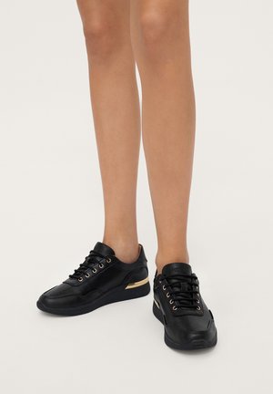 Piernas con zapatillas deportivas bajas negras con ojales dorados y un detalle dorado en el talón, de pie sobre un suelo blanco.