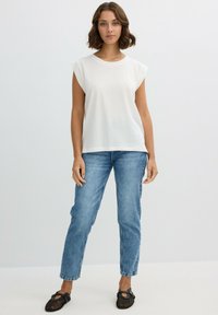 Top sin mangas de algodón blanco con un escote redondo, combinado con unos jeans de mezclilla azul ligeramente holgados y zapatos planos negros.