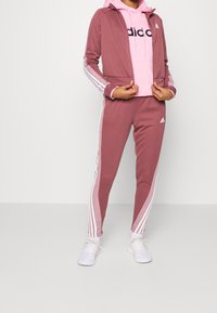 Różowy dres składający się z zapinanej kurtki z kontrastującymi białymi paskami i logo, w komplecie z dopasowanymi spodniami i białymi sneakersami.