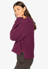 Strickpullover in tiefem Lila mit gerippter Textur, lockerer Passform, seitlichen Schlitzen und herunterhängenden Schultern. Kombiniert mit dunklen Hosen.