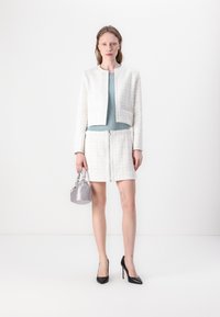 Witte tweedjas met een cropped ontwerp, gecombineerd met een lichtblauwe sweater en een bijpassende rok met rits aan de voorkant; zwarte hoge hakken en een glanzende zilveren tas.