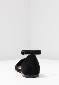 Ballerines en suède noir avec une bride autour de la cheville, un bout pointu et une semelle plate en caoutchouc. Texture lisse sans motifs visibles.