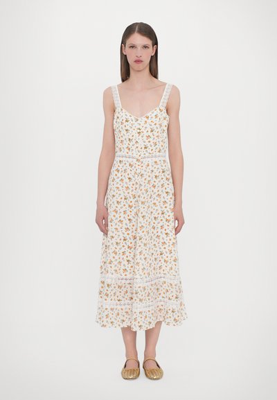 Robe florale blanche avec des fleurs orange, des bretelles en dentelle et un ourlet en volants. Fini avec un accent en dentelle à la taille. Portée avec des chaussures en osier dorées.