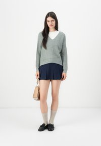 Grønn strikket v-hals genser med ribbestruktur, marineblå plissert miniskjørt, beige knehøye sokker og svarte loafers, holder en beige håndveske.