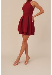 Nainen, jolla on yllään hihaton, kerroksellinen burgundinvärinen mekko, jossa on poimutuksia, yhdistettynä kultaisiin nauhakorkokenkiin ja vähäisiin koruihin.