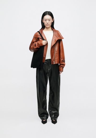 Casaco de couro castanho com colarinho grande, suéter creme, calças de couro pretas e sapatos de salto pretos. O modelo segura uma mala tote preta.