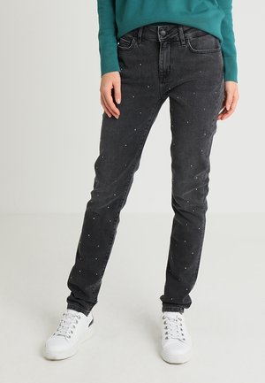 Persoon draagt zwarte slim-fit jeans met kleine decoratieve studs, een teal lange mouwen top en witte sneakers, staand op een effen achtergrond.