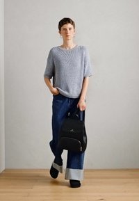 Maglione grigio chiaro a maglia con maniche corte, abbinato a jeans a gamba larga di un blu scuro e uno zaino nero, su uno sfondo di una parete neutra e un pavimento in legno.