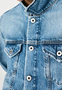 Detailní záběr na světle modrou džínovou bundu s kovovými knoflíky a náprsními kapsami, nošenou s límečkem vztyčeným.