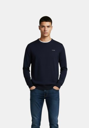 Jonge man met een donkerblauwe trui over een wit overhemd en blauwe spijkerbroek, staand met handen in de zakken tegen een effen achtergrond.