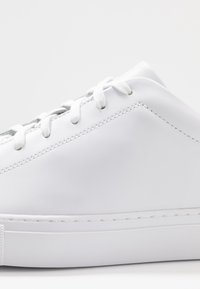 Vit sneakers i läder med slät yta, rund tå, vita snören och enkel design, med minimal sömnad längs sulan.