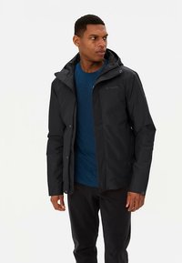 Zwarte waterdichte jas met een afneembare capuchon, drukknopen aan de voorkant en zakken met rits. Draagt over een blauw T-shirt en zwarte broek.