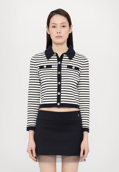 Gilet tricot rayé avec des rayures horizontales bleu marine et blanches, doté d'un col, d'une fermeture à boutons et de deux poches poitrine. Associé à une jupe noire.