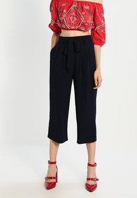 Marinblå cropped byxor med knytmidja, med två sidofickor och en mjuk textur. Matchas med en röd broderad crop top och röda klackar.