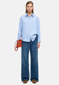 Kobieta w jasnoniebieskiej w paski koszuli, w szerokich niebieskich dżinsach, w brązowych butach, trzymająca małą rudawą torebkę na tle białym.