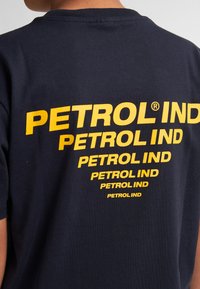 Černé bavlněné tričko s krátkým rukávem a kulatým výstřihem, na zadní straně zdobené velkým žlutým nápisem "PETROL IND", který je vytištěn několikrát.