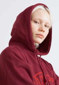 Μαραoon φούτερ με κορδόνι και μεγάλη μπροστινή τσέπη. Μαλακή υφή, με έντονα πορτοκαλί γράμματα στο στήθος και χαλαρή εφαρμογή.