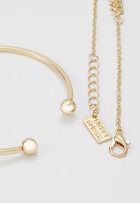 Bracelet en métal doré avec un design circulaire lisse, comprenant deux accents sphériques et une chaîne liée, se terminant par un fermoir en homard et une étiquette.
