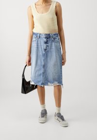 Falda de mezclilla azul clara con dobladillo deshilachado, combinada con una blusa de punto beige y zapatillas grises. Bolso negro sostenido en la mano derecha.