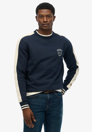 Marineblauwe katoenen sweatshirt met crèmekleurige accenten en gestreepte manchetten, voorzien van een klein logo op de borst. Geverfde in combinatie met blauwe denim jeans.