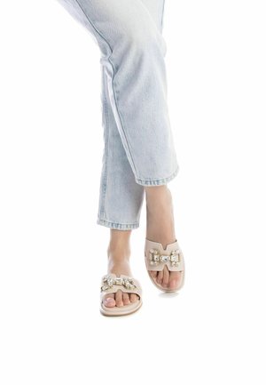 Sandalias beige sin respaldo con una banda ancha que presenta una cadena decorativa y acentos de strass. Material suave, suela plana y diseño de punta abierta.
