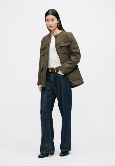 Bruine en tan geruite oversized jas met twee voorzakken, gedragen over een witte shirt, gecombineerd met donkerblauwe wijde jeans en zwarte schoenen.