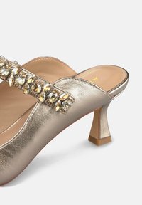 Mules metálicos dorados con tacón, con punta redondeada, que presentan una tira decorativa de strass y un tacón de bloque de altura media. Textura suave.