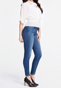 Weiße, halbtransparente Bluse mit fransigen Ärmeln, kombiniert mit dunkelblauen, engen Jeans und schwarzen, spitzen High Heels. Glattes Material und taillierte Passform.