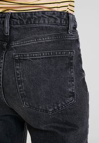 Jeans de denim negro que muestran un primer plano de la parte trasera, con un bolsillo visible y costuras prominentes. El material es suave y tiene un ligero brillo.