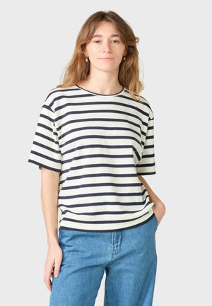 Młoda kobieta o jasnobrązowych włosach, ubrana w biało-granatową koszulkę w paski z krótkim rękawem i niebieskie dżinsy, stojąca z jedną ręką w kieszeni.