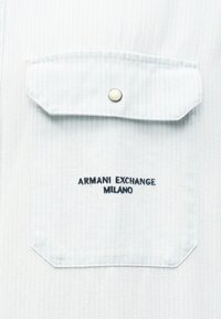 Φωτεινή μπλε τσεπάκι από τζιν με καπάκι και μεταλλικό κουμπί, με μαύρο κεντητό κείμενο "ARMANI EXCHANGE MILANO".