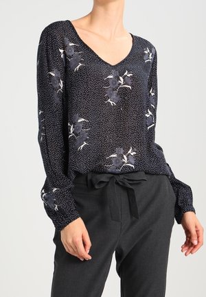 Blusa nera floreale con scollatura a V, maniche lunghe e motivo a pois bianchi. Abbinata a pantaloni grigi con dettagli in vita a nastro.