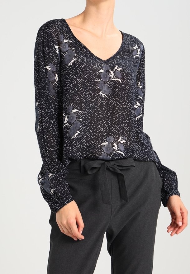 Blusa nera floreale con scollatura a V, maniche lunghe e motivo a pois bianchi. Abbinata a pantaloni grigi con dettagli in vita a nastro.