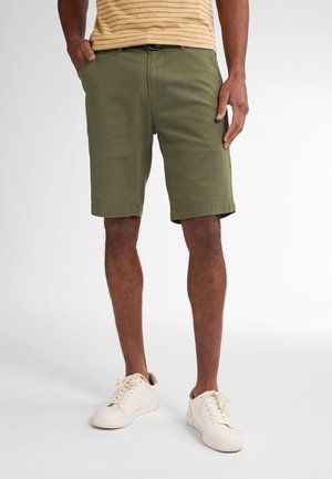 Grüne Baumwollshorts mit strukturiertem Muster, seitlichen Taschen und umgeschlagenem Saum, kombiniert mit weißen Sneakers.