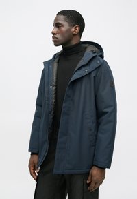 Muž v námornícko modrej zimnej bunde s kapucňou a čiernym rolákom, stojaci bokom pred jednoduchým svetlým pozadím.