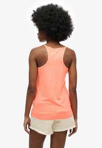 Canotta arancione neon con design a schiena scoperta, realizzata in tessuto leggero. Presenta un orlo dritto e una vestibilità comoda. Abbinata a pantaloncini color crema.