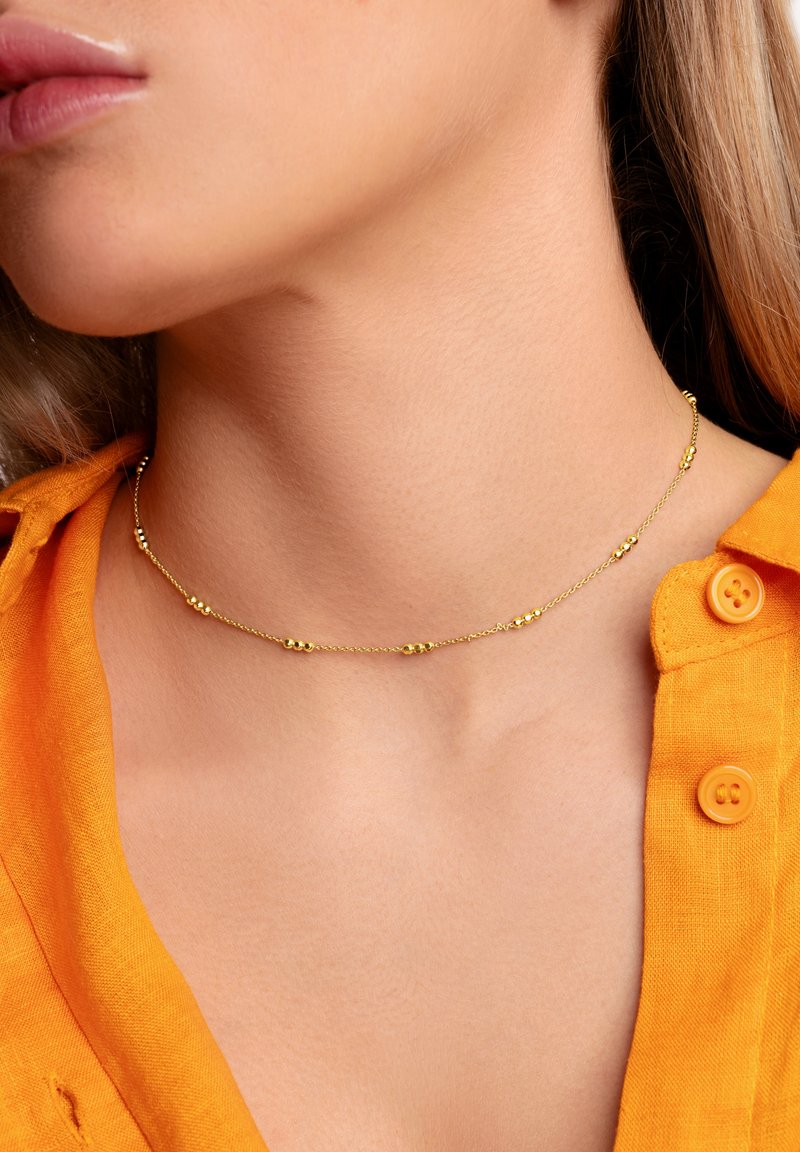 Collar de cadena de oro con pequeñas cuentas redondas, llevado sobre un cuello con piel suave, combinado con una camisa de botones naranja.