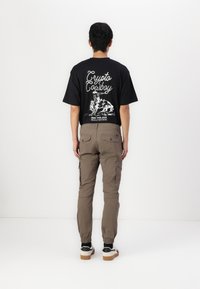 Camiseta negra con gráfico blanco de "Crypto Cowboy" en la parte posterior, combinada con pantalones cargo beige con bolsillos laterales y calzado casual.