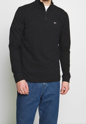 Czarny długi sweter z długim rękawem i suwakiem do połowy długości, wykonany z gładkiego materiału, z małym logo na lewej piersi i ściągaczami na mankietach.