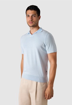 Ung mand med kort mørkt hår iført en lyseblå poloshirt med korte ærmer og beige bukser mod en ensfarvet baggrund.