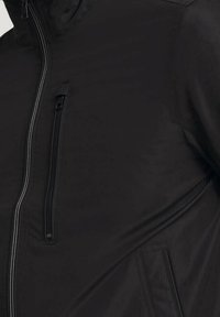 Černá bunda s texturovaným povrchem, vybavená vertikální kapsou na hrudi na zip a hladkým designem. Viditelné minimalistické a funkční detaily.