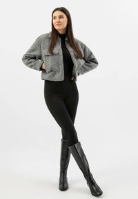 Šedá zkrácená bunda se dvěma předními kapsami, nošena přes černý top a legíny, v kombinaci s černými vysokými botami až ke kolenům. Jednoduché bílé pozadí.